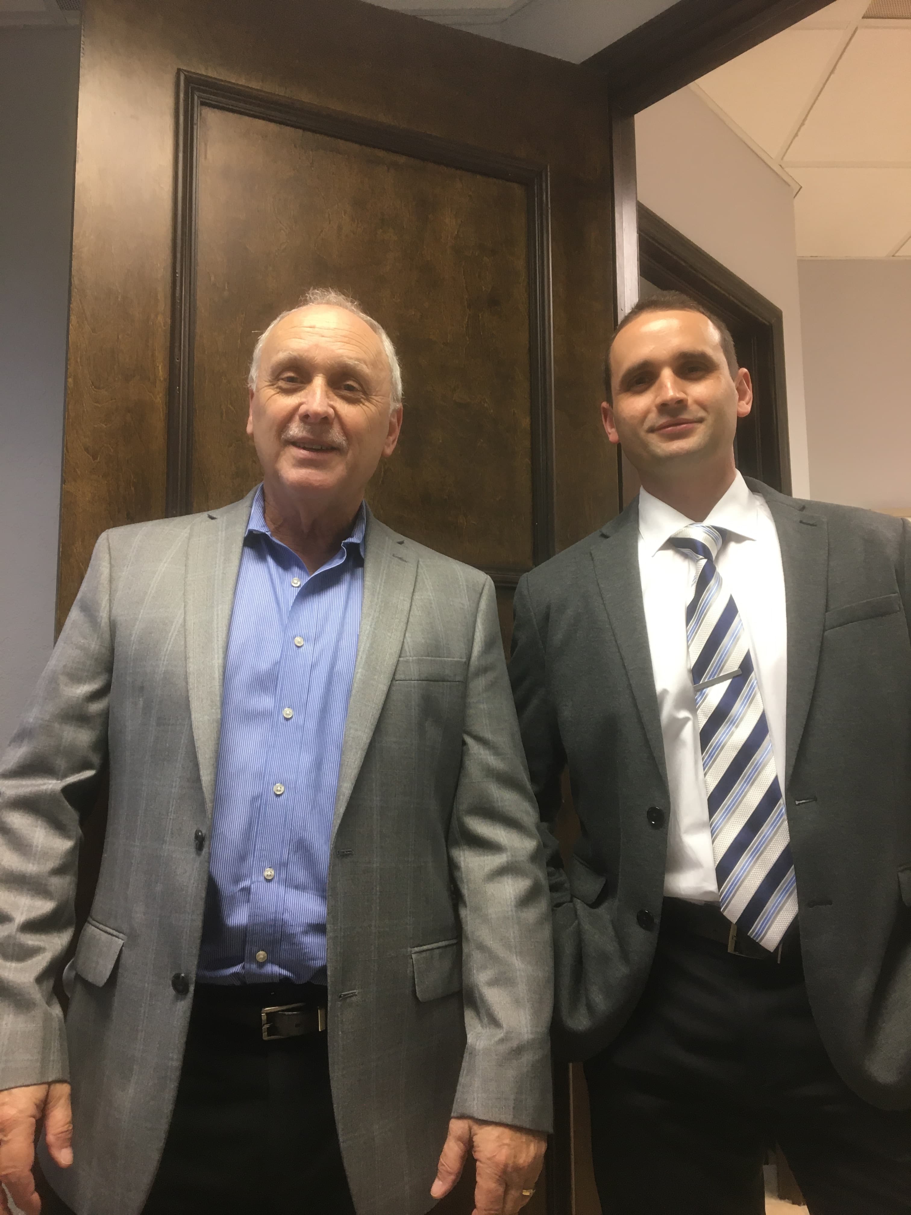 Younger man in dark suit and older man in light grey suit posing indoors.
