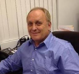 Smiling middle-aged man with light hair wearing a blue button-down shirt in an office.