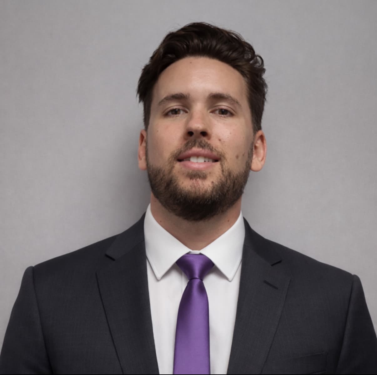 Professional headshot of a smiling man with a beard wearing a suit and purple tie.
