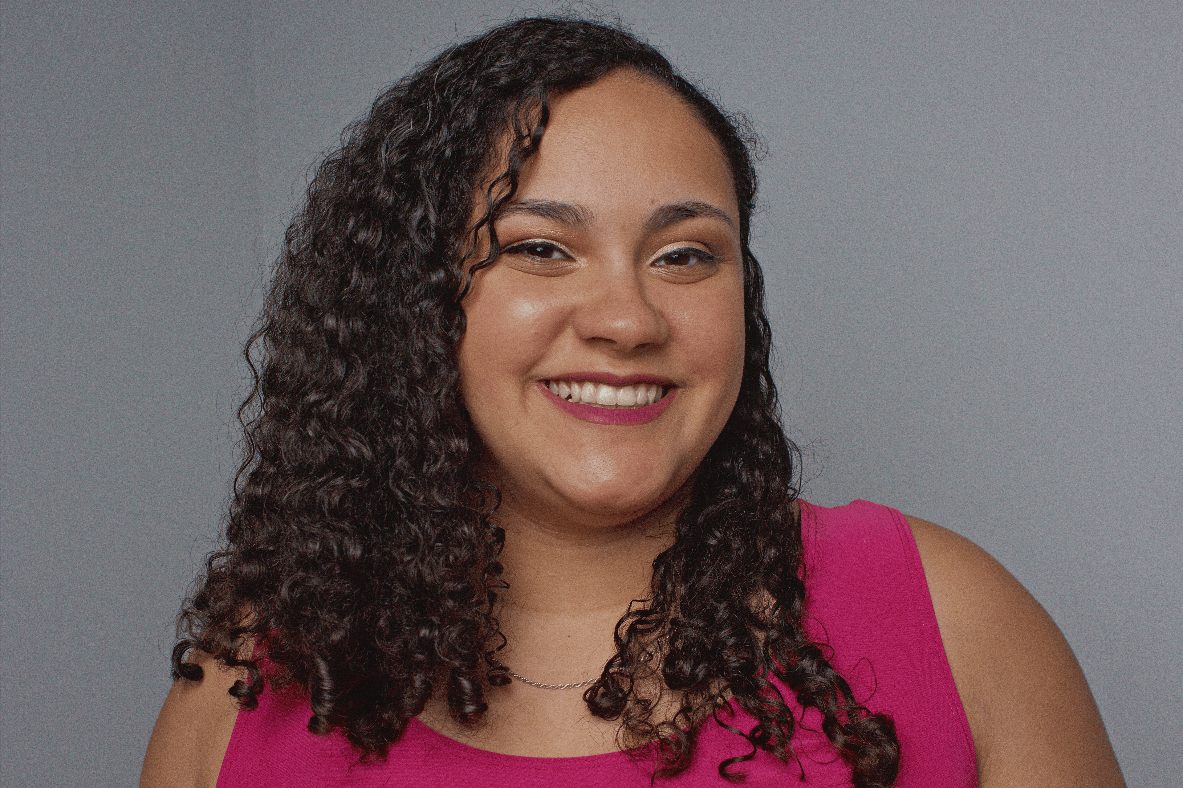 Smiling woman with long dark curly hair wearing a pink top against gray background.
