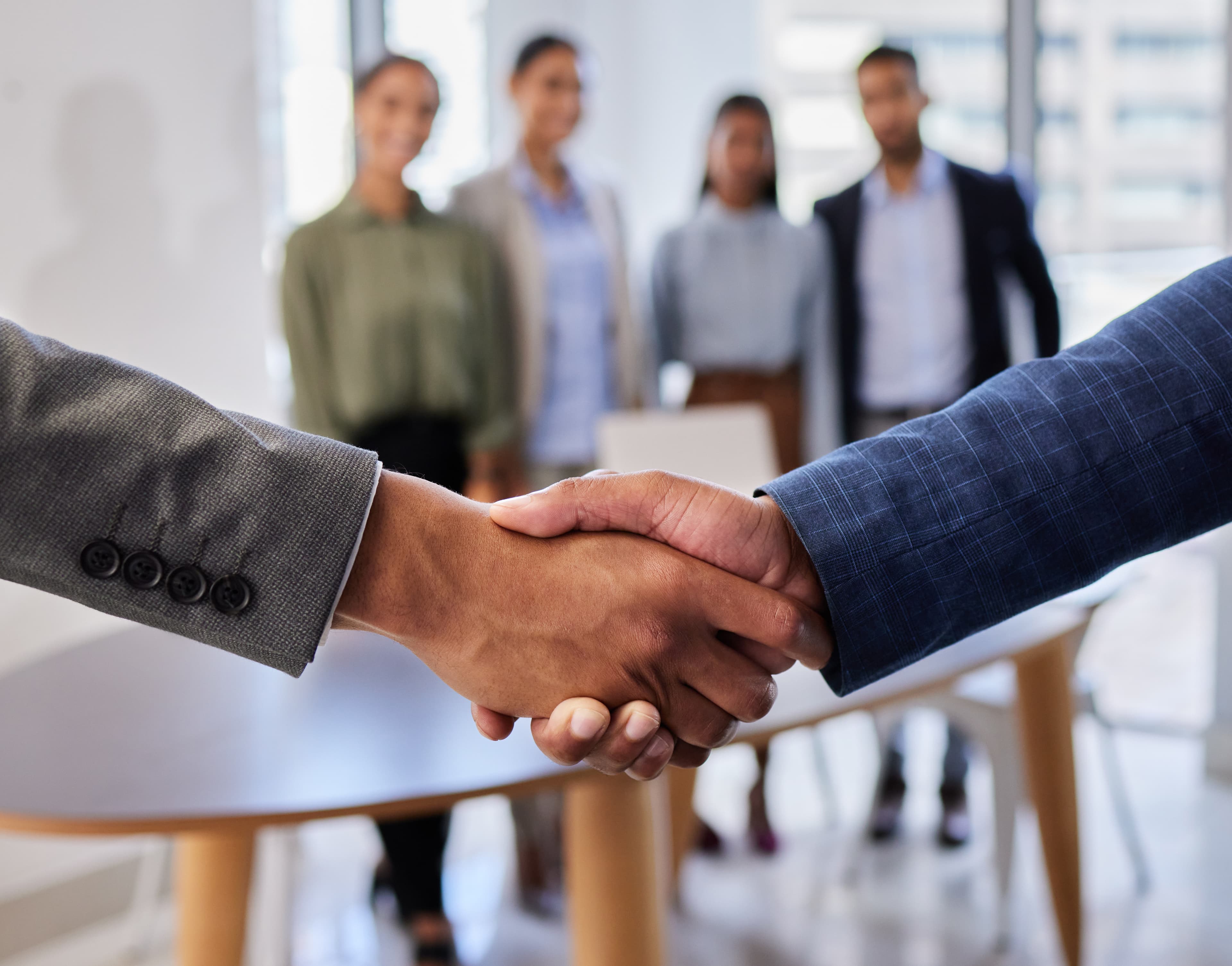 Two business professionals shaking hands with a diverse, smiling team in the blurred background.