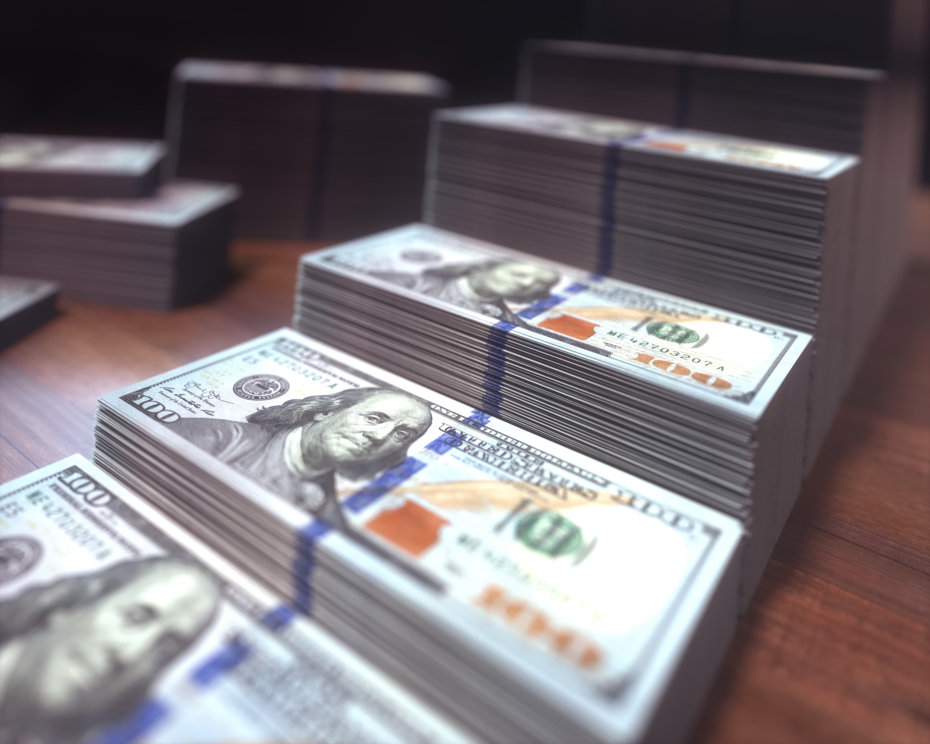 Multiple large stacks of US hundred-dollar bills arranged on a dark wooden surface.